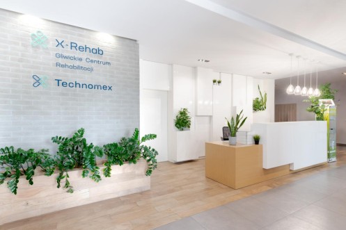Recepcja X-Rehab Gliwice z logo na ścianie, jasnym wnętrzem i zielonymi roślinami w strefie wejściowej. 