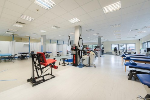 Przestronna sala terapii z profesjonalnymi maszynami Keiser i stanowiskami do rehabilitacji. 