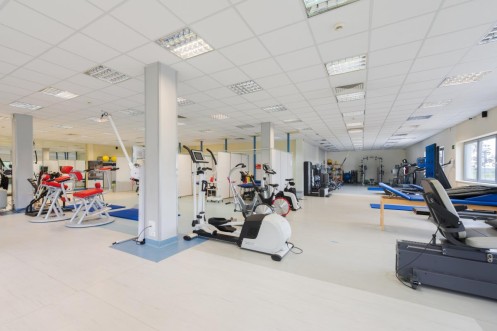 Przestronna sala rehabilitacyjna z rowerami treningowymi i stołami do terapii manualnej. 
